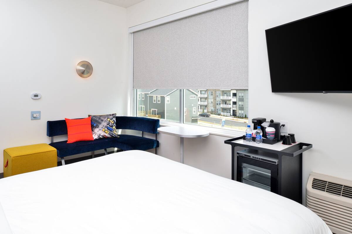 A hotel room with a bed, a blue sofa with red cushions, a small table with a coffee maker, and a window with a view of buildings.