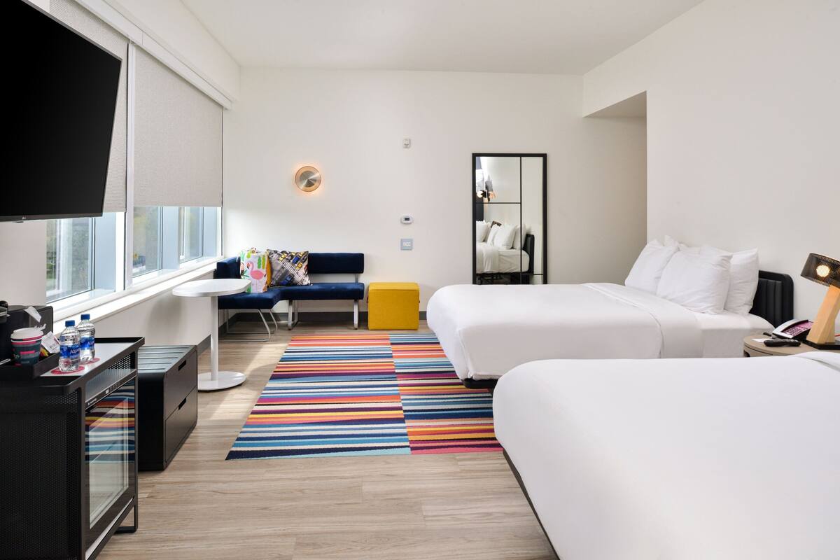 A modern hotel room with a large bed, a smaller bed, a desk with a chair, a colorful striped rug, and a large window with blinds.