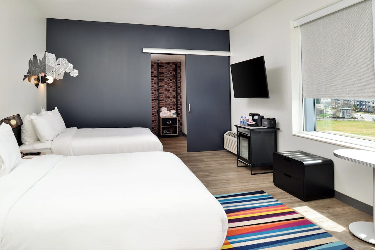 A modern hotel room with two beds, a flat-screen TV, a desk with a computer, and a window with a city view.