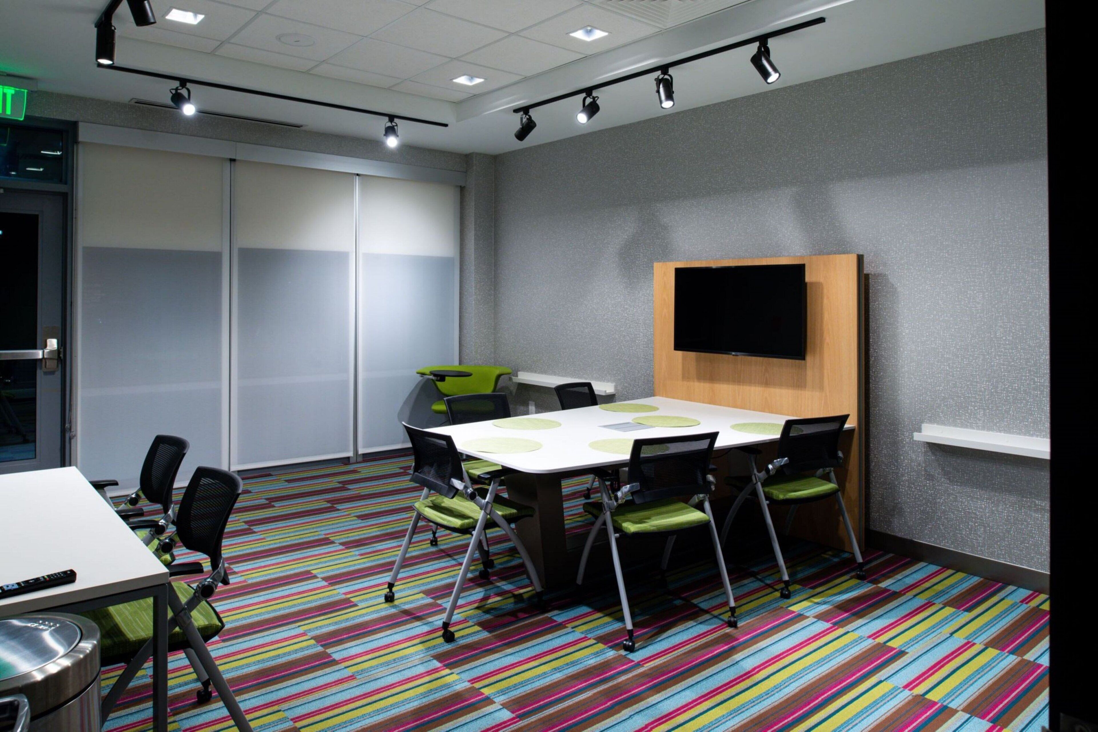 A conference room with a large table, chairs, a flat-screen TV, and a colorful striped carpet.