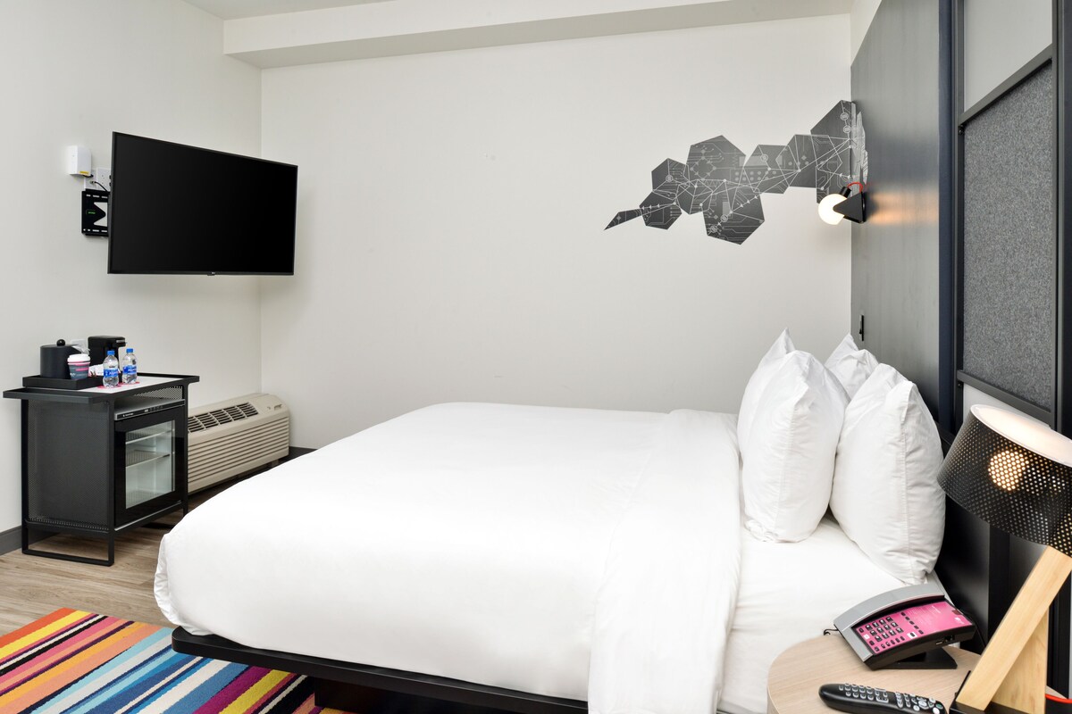 Habitación de hotel con cama, televisor de pantalla plana en la pared, mesita de noche con lámpara y una alfombra a rayas colorida.