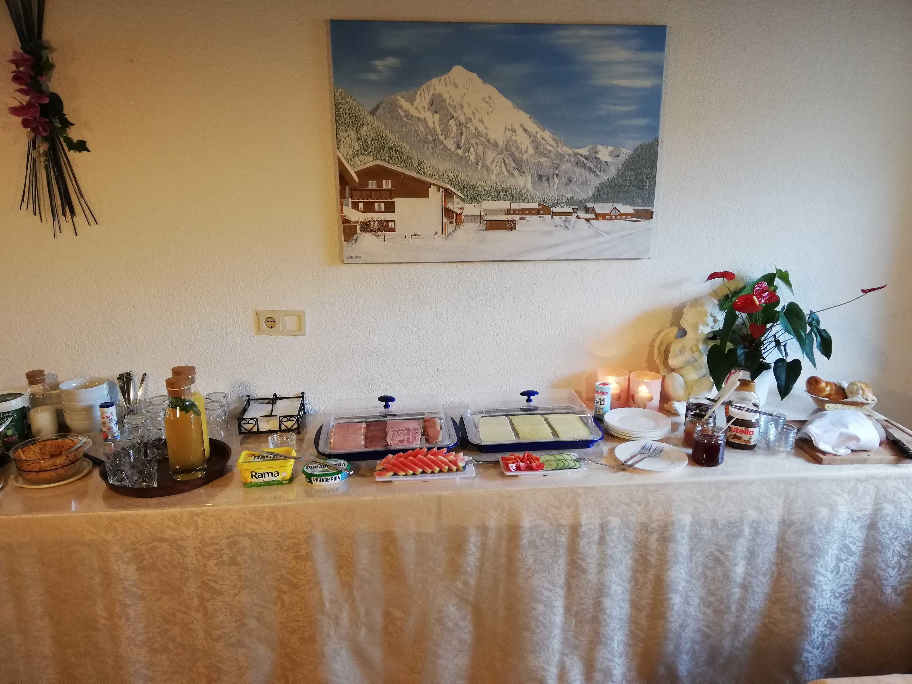 Une table de buffet garnie de divers plats et condiments, un tableau représentant un paysage de montagne et une plante aux fleurs rouges.