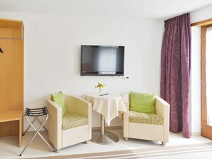Une pièce comprenant deux fauteuils, une petite table avec un vase, un téléviseur fixé au mur et une fenêtre ornée de rideaux violets.