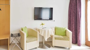 Une pièce comprenant deux fauteuils, une petite table avec un vase, un téléviseur fixé au mur et une fenêtre ornée de rideaux violets.