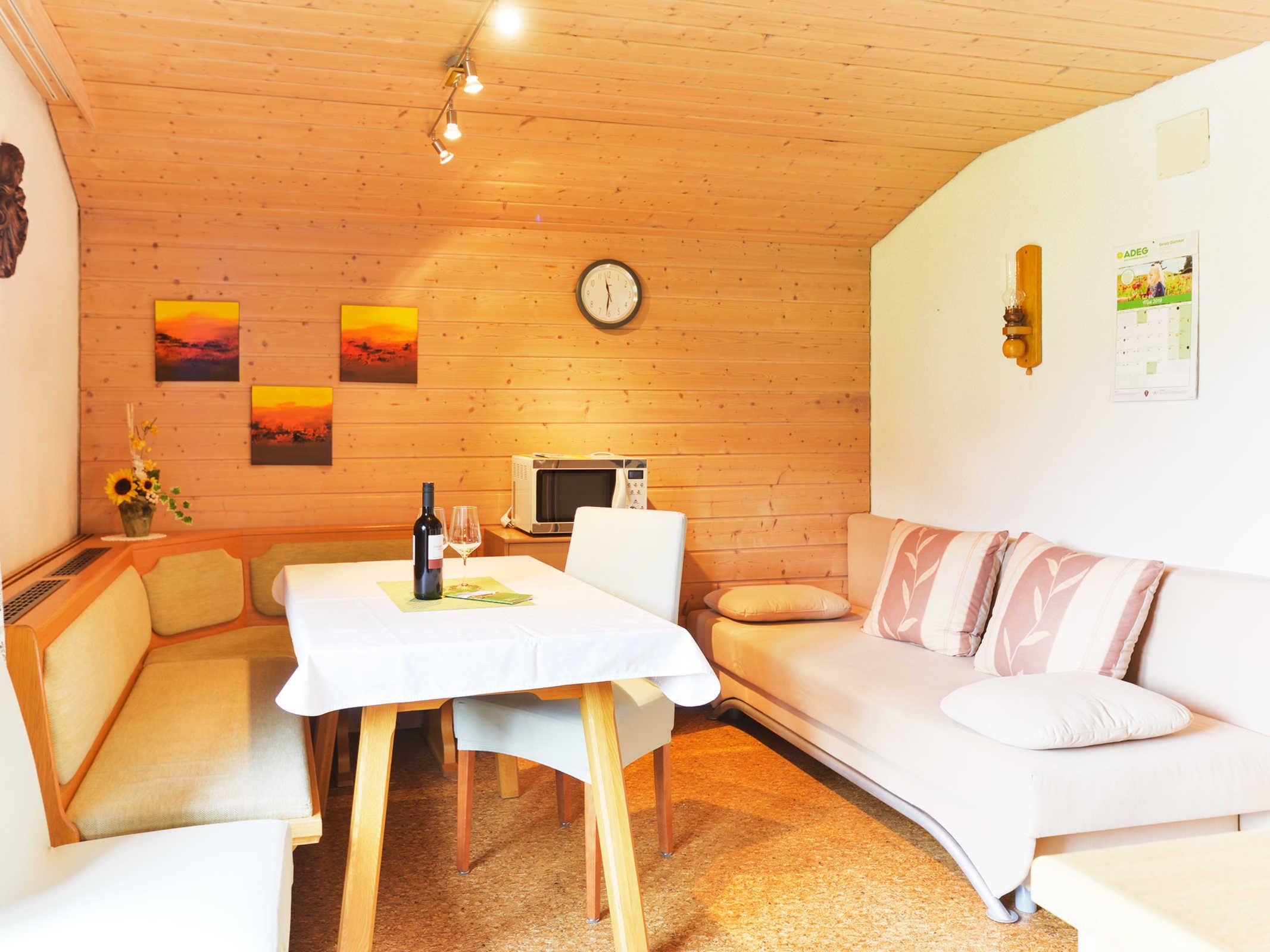 A cozy room with a wooden ceiling, a white sofa, a dining table with a wine bottle and glasses, and a clock on the wall.
