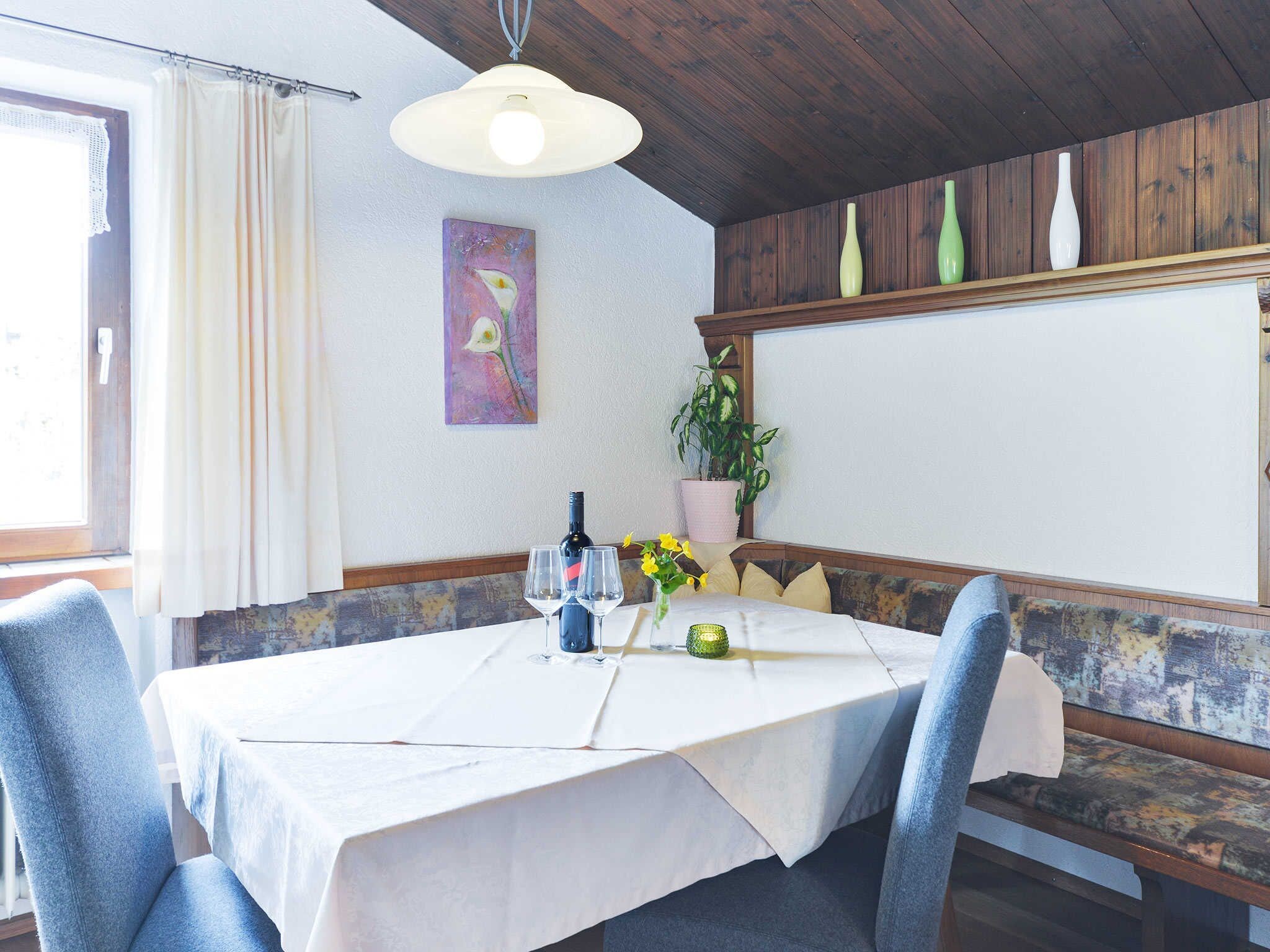 A dining area with a table set for two, a potted plant, and a painting on the wall.