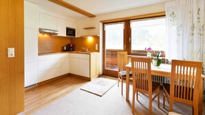 Een compacte keuken met een magnetron, spoelbak en houten keukenkastjes. Er is een eethoek met een tafel en stoelen, en een raam met gordijnen.