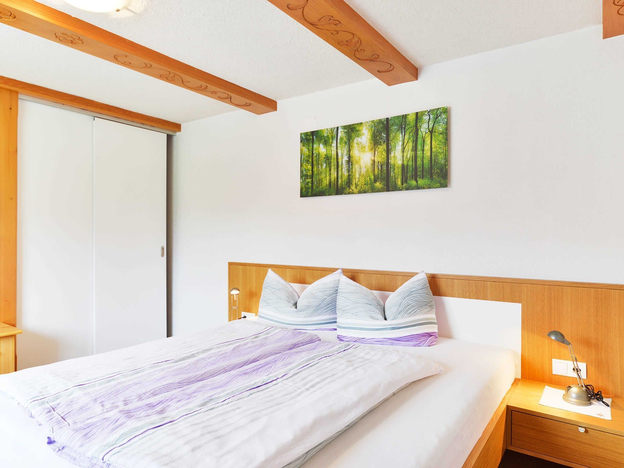 Een slaapkamer met een bed, nachtkastje, lamp, een boslandschap op canvas en houten balken in het plafond.