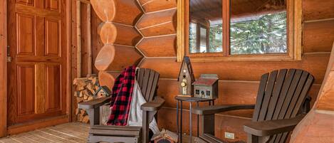 A log cabin interior with a wooden door, two Adirondack chairs, a small table with a radio, and a window overlooking trees.