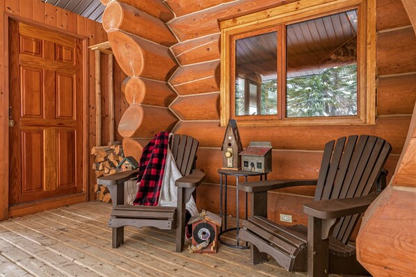 Interior de una cabaña de troncos con una puerta de madera, dos sillas Adirondack, una mesa pequeña con una radio y una ventana con vistas a árboles.