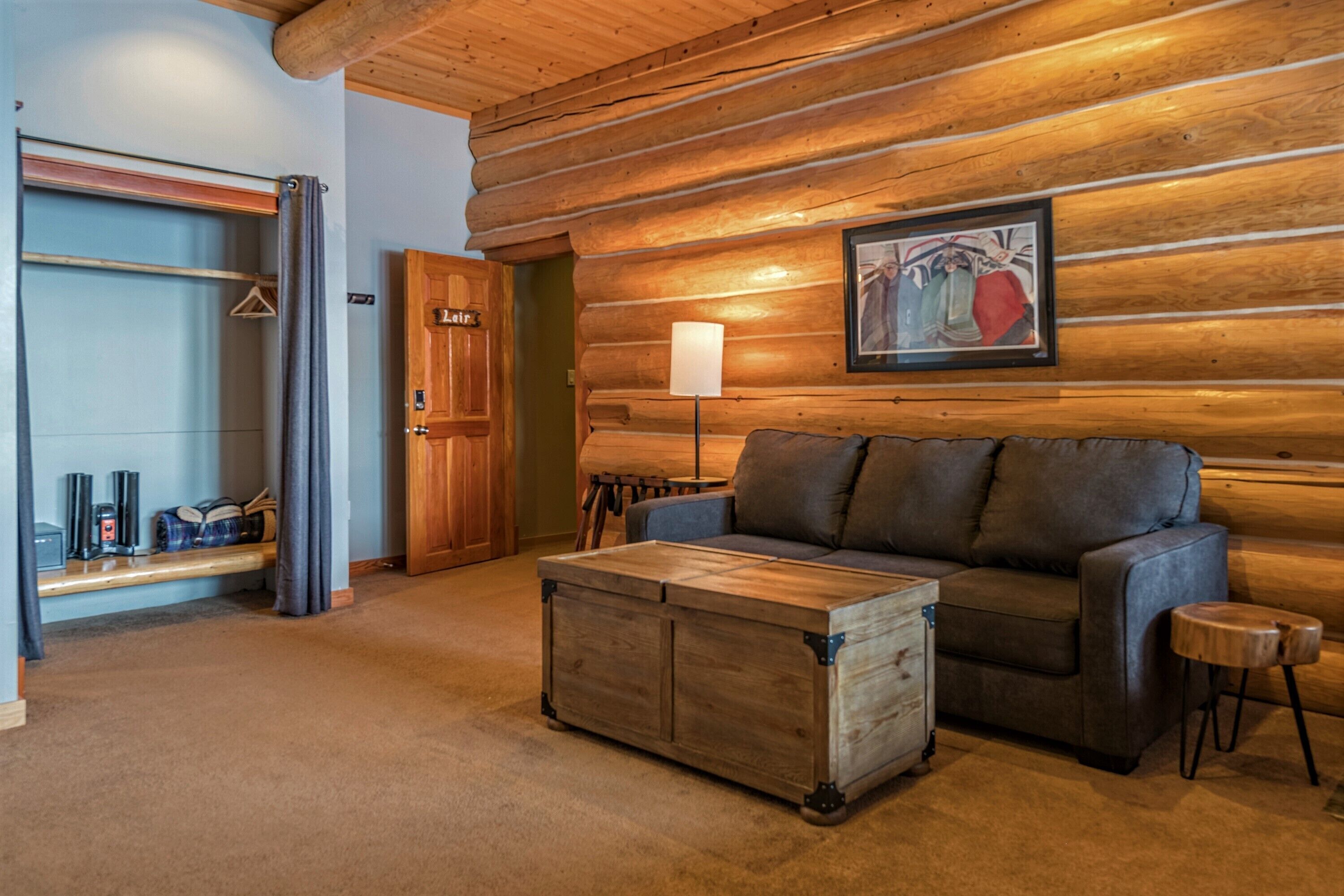 A log cabin interior with a sofa, a wooden chest, a lamp, and a coat rack.