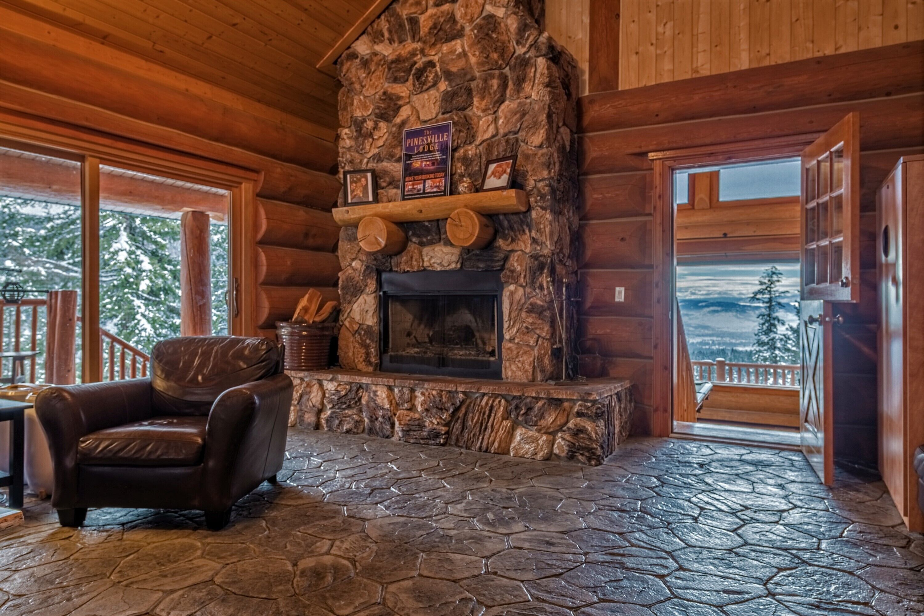 A log cabin interior with a stone fireplace, leather chair, and a view of the outdoors.
