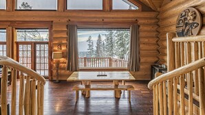 A wooden cabin interior with a large wooden table and benches, a view of trees through large windows, and a wooden railing. - Spirit Lodge at Silverstar (Vernon)