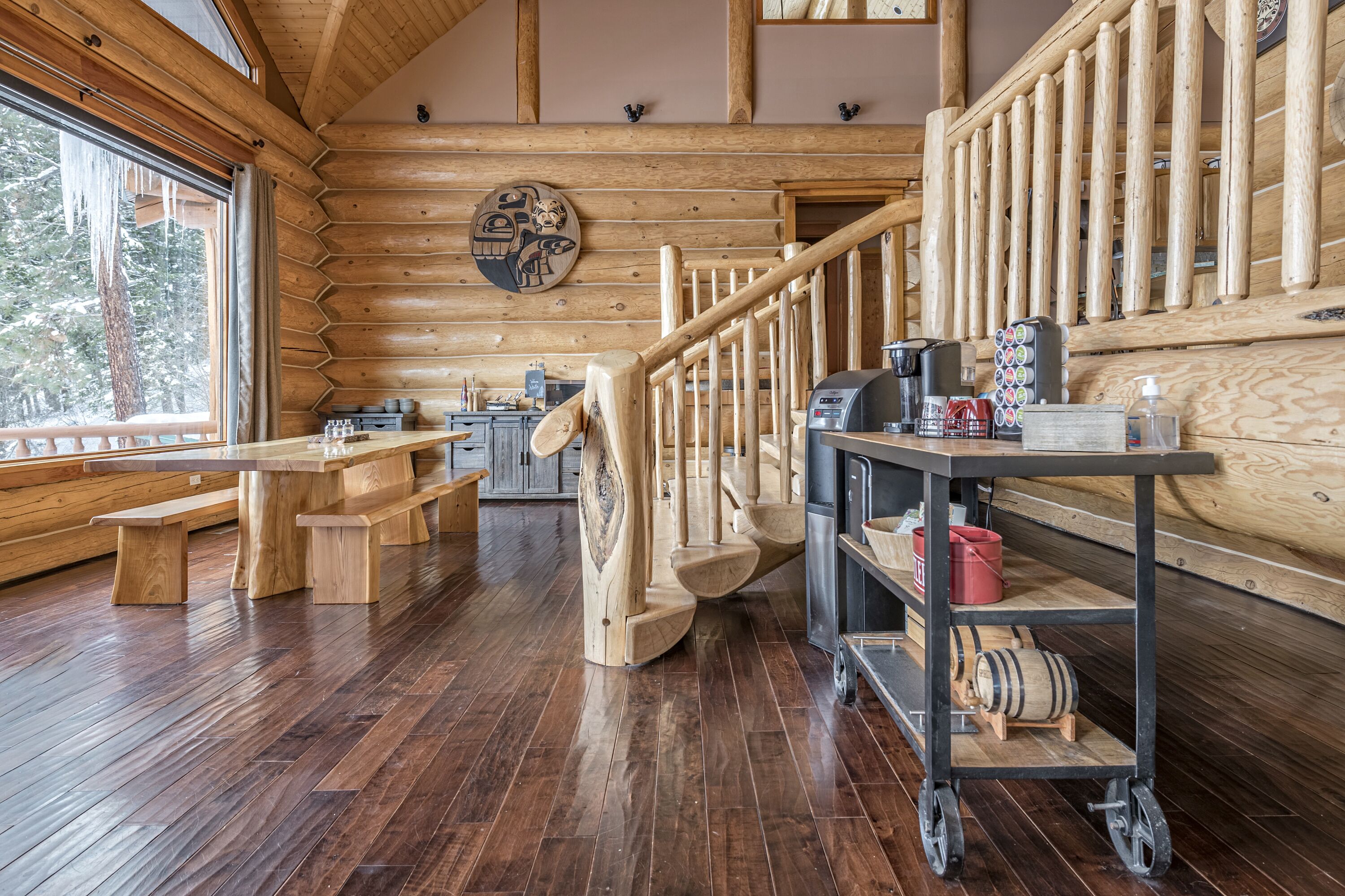 A log cabin interior with wooden floors, a staircase, wooden benches, a coffee station, and a wooden wall with a circular artwork.