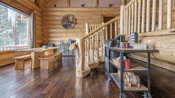 A log cabin interior with wooden floors, a staircase, wooden benches, a coffee station, and a wooden wall with a circular artwork.