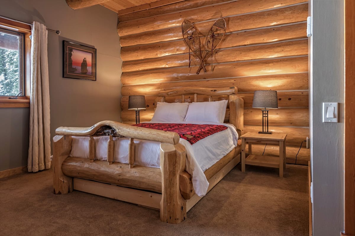 A log cabin bedroom with a wooden bed, two bedside tables, a lamp, and a framed picture on the wall.