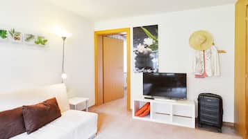 Una stanza moderna e luminosa, con un divano bianco, un televisore a schermo piatto, un piccolo tavolino bianco, una lampada, una valigia e una porta in legno che dà su un'altra stanza.