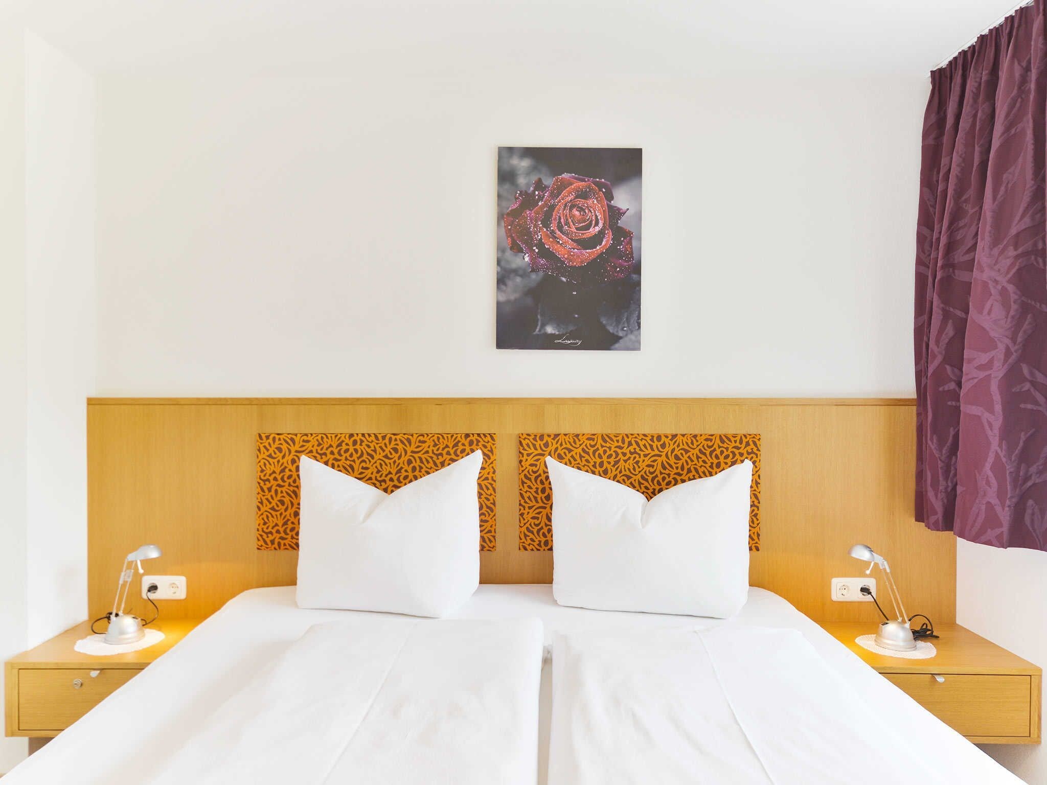 A double bed with white linens, a wooden headboard with a patterned design, two bedside tables with lamps, and a framed artwork of a rose on the wall.