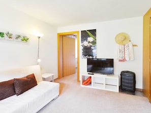 A modern, well-lit room with a white sofa, a flat-screen TV, a small white side table, a lamp, a suitcase, and a wooden door leading to another room. - Apartment Toblermann - Pension Oberhauser (Schoppernau)