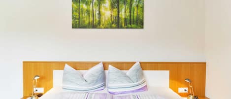 Habitación de hotel con cama, dos mesitas de noche, lámpara de noche, cuadro colgado en la pared que representa un bosque y vigas de madera en el techo.