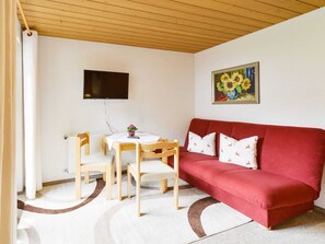 A room with a red sofa, a TV mounted on the wall, a small table with chairs, and a painting of sunflowers. - Double room 2nd floor - Pension Oberhauser (Schoppernau)