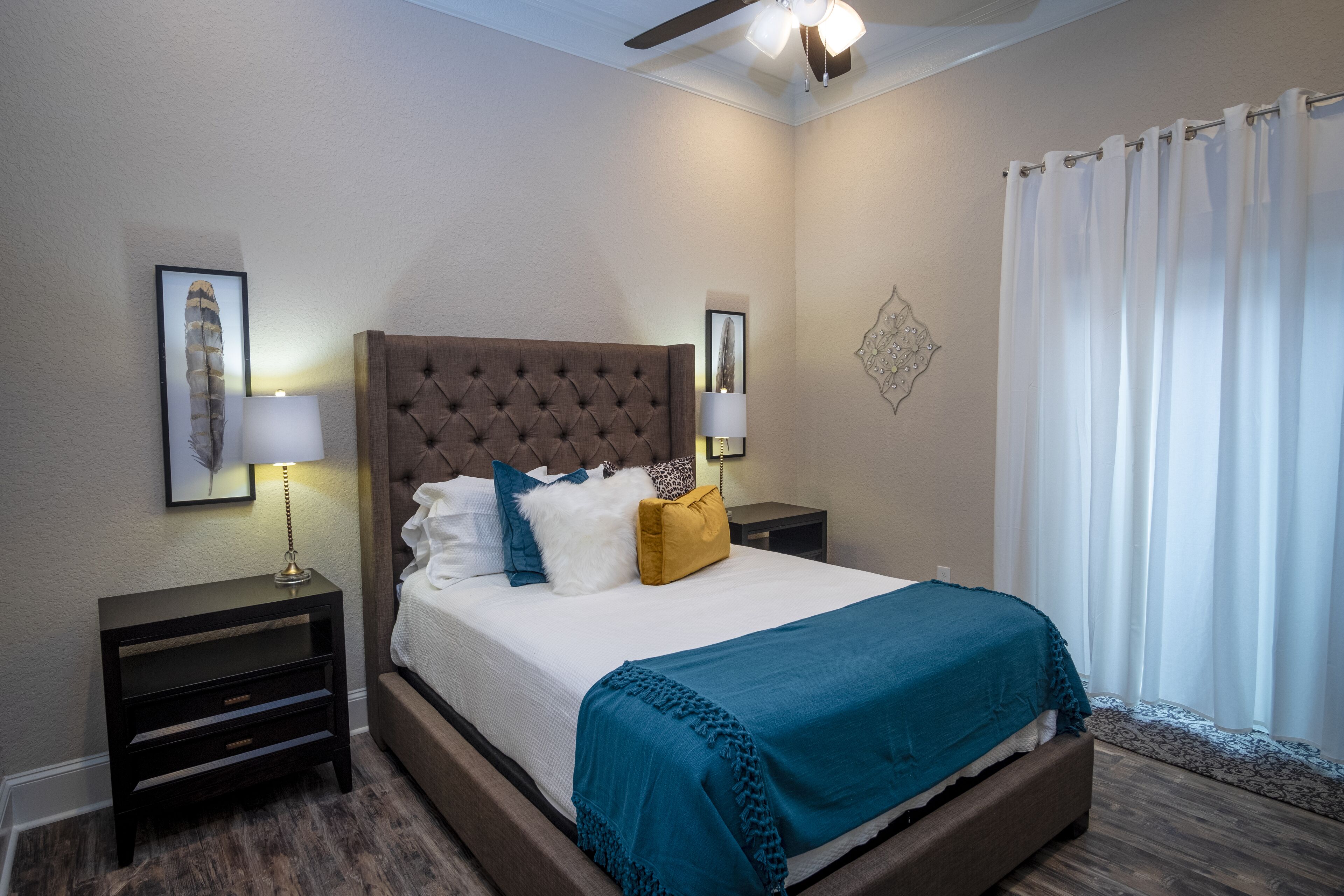 Un dormitorio con cabecera capitonada, cama con ropa de cama blanca y turquesa, dos mesitas de noche con lámparas y un ventanal con cortinas.