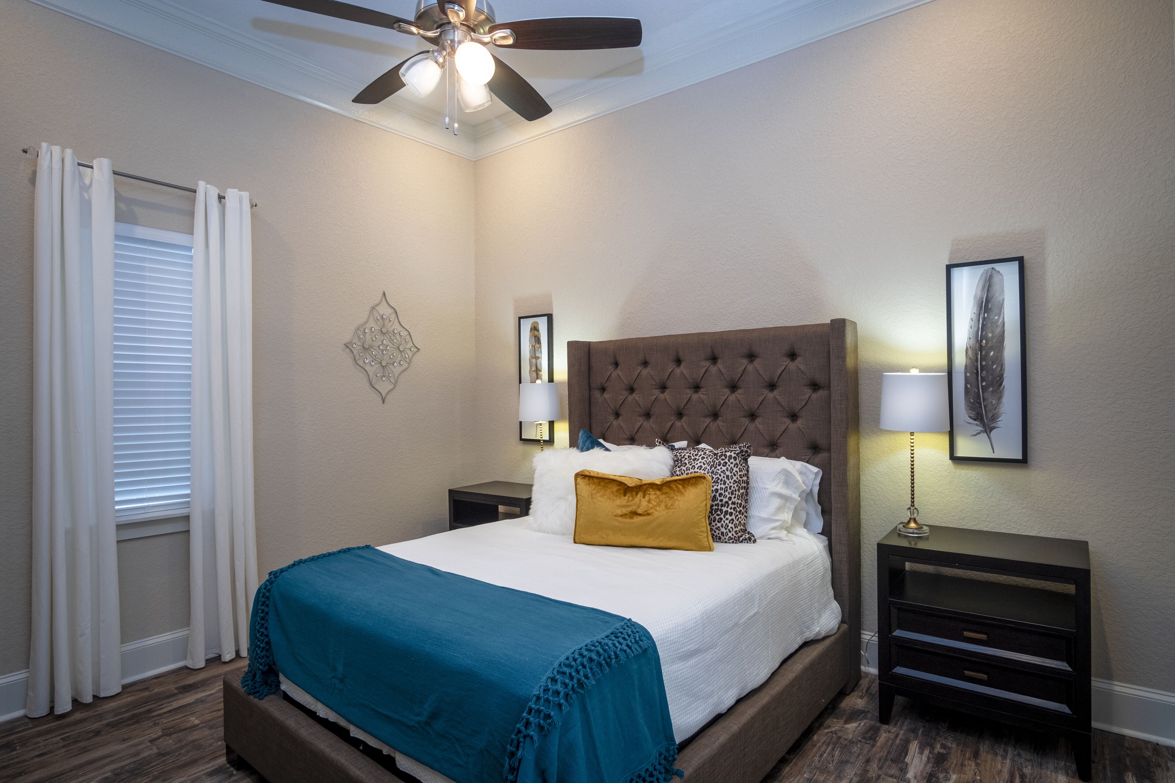 Quarto com cabeceira estofada, cama com manta azul-turquesa, duas mesas de cabeceira com abajures, um ventilador de teto e uma janela com cortinas brancas.