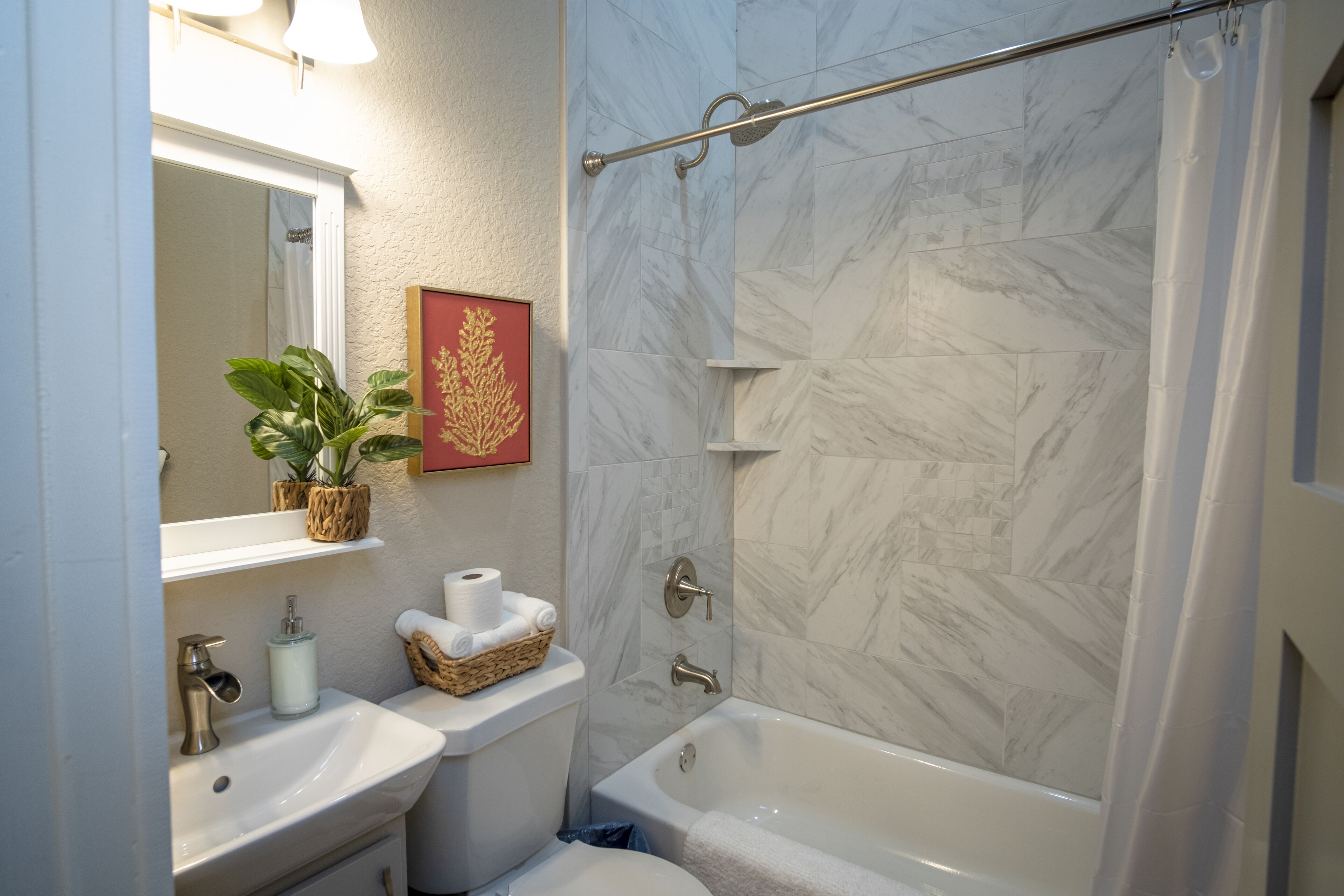 A bathroom with a marble shower wall, a bathtub, a sink, a toilet, and a potted plant.