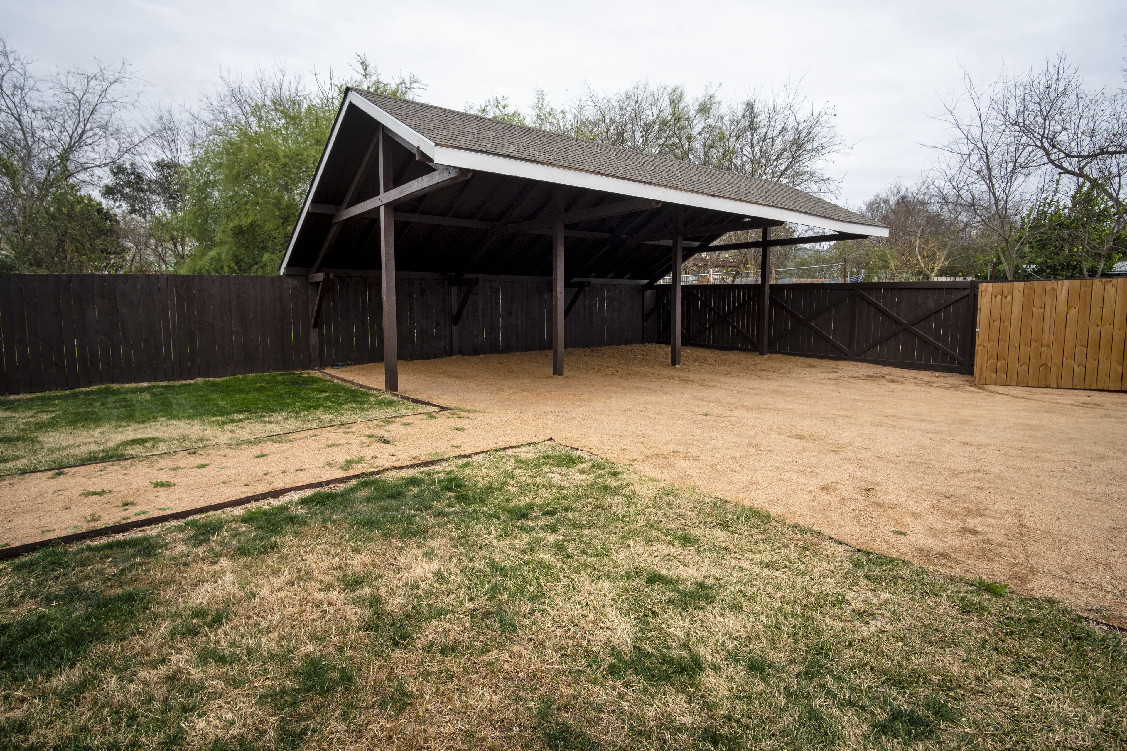 Une étable dotée d’un toit en métal et de murs en bois, entourée d’une zone délimitée par une clôture, où l’on trouve un mélange d’herbe et de terre.