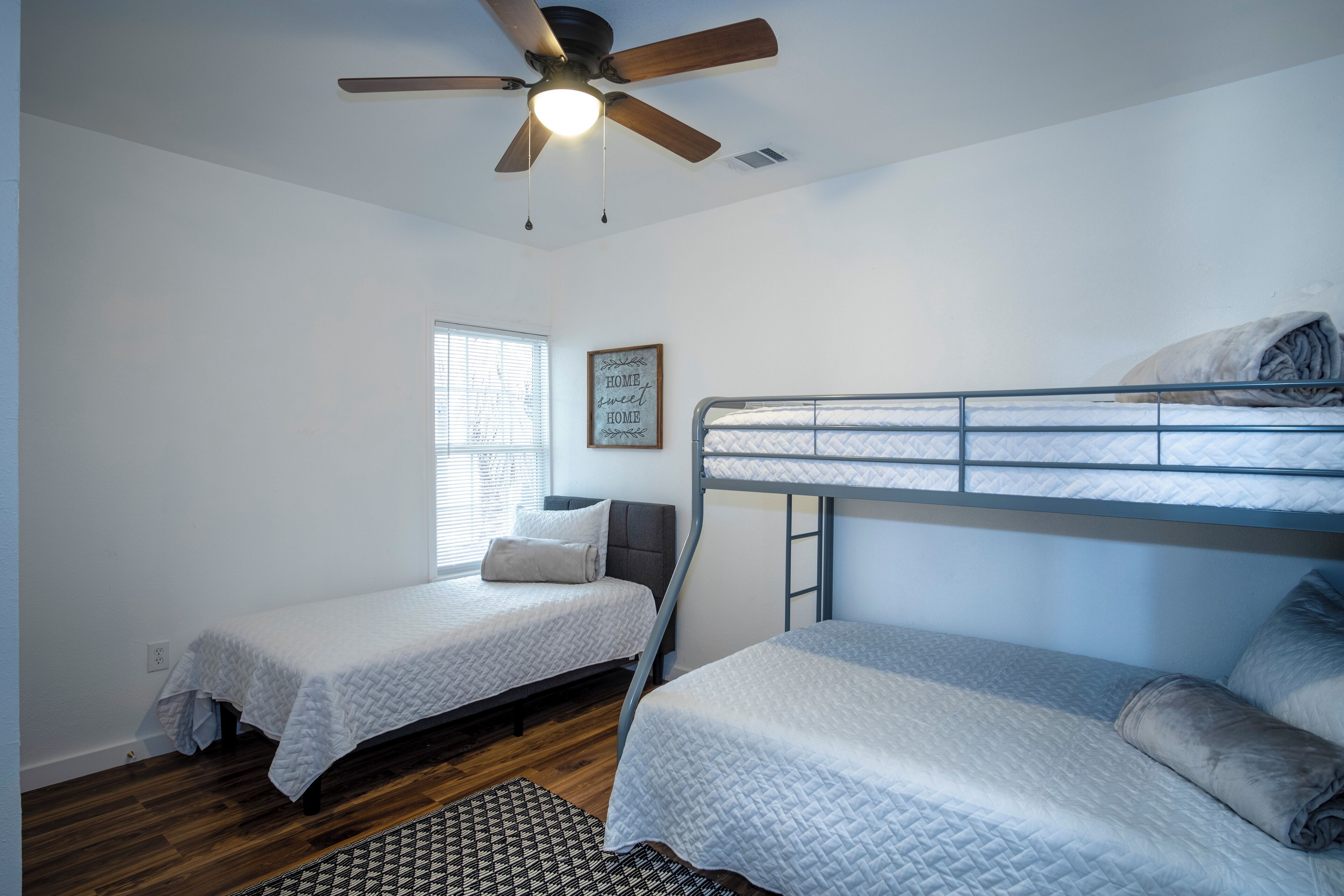 A bedroom with two single beds, a ceiling fan, a window with blinds, and a framed sign on the wall.