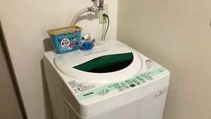 A front-loading washing machine with a control panel, a water inlet hose, and a detergent container on top. - Espoir (Kumamoto)