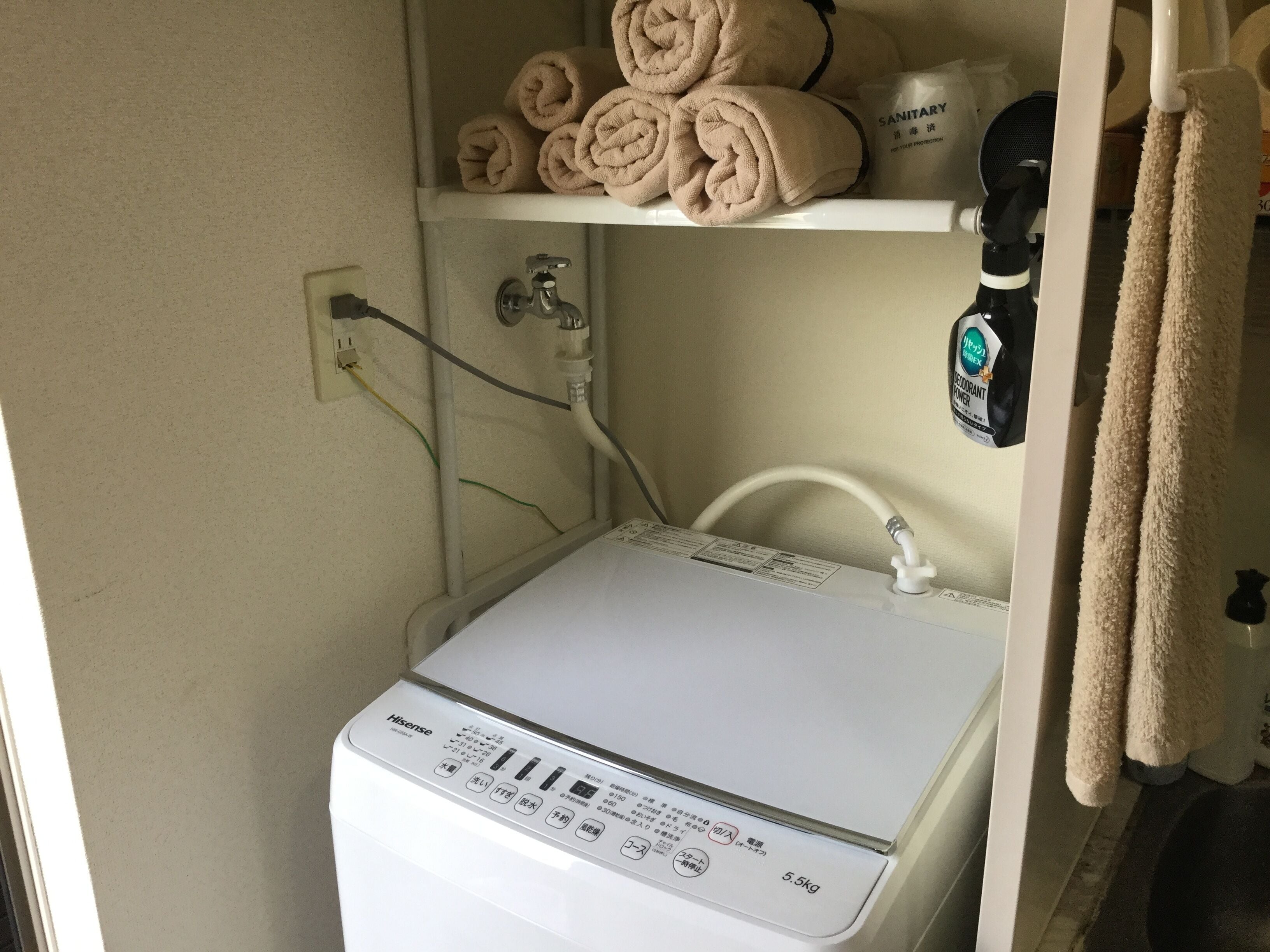 A compact, white Hisense washing machine with a control panel, located in a small laundry room with a shelf above holding rolled towels and a bag labeled "SANITARY".