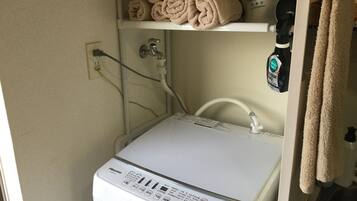 A compact, white Hisense washing machine with a control panel, located in a small laundry room with a shelf above holding rolled towels and a bag labeled "SANITARY".