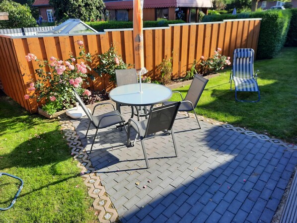 A patio with a round table and chairs, surrounded by a wooden fence and a garden with pink flowers.