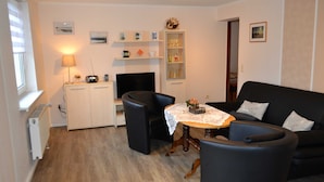 A living room with a black sofa, a small round table with a lace tablecloth, a TV, and shelves with decorative items. - Wendt apartment, Germany (Insel Poel)