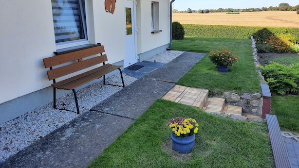 A white building with a brown bench outside, a satellite dish, and a well-maintained garden with flowers and a lawn. - Wendt apartment, Germany (Insel Poel)