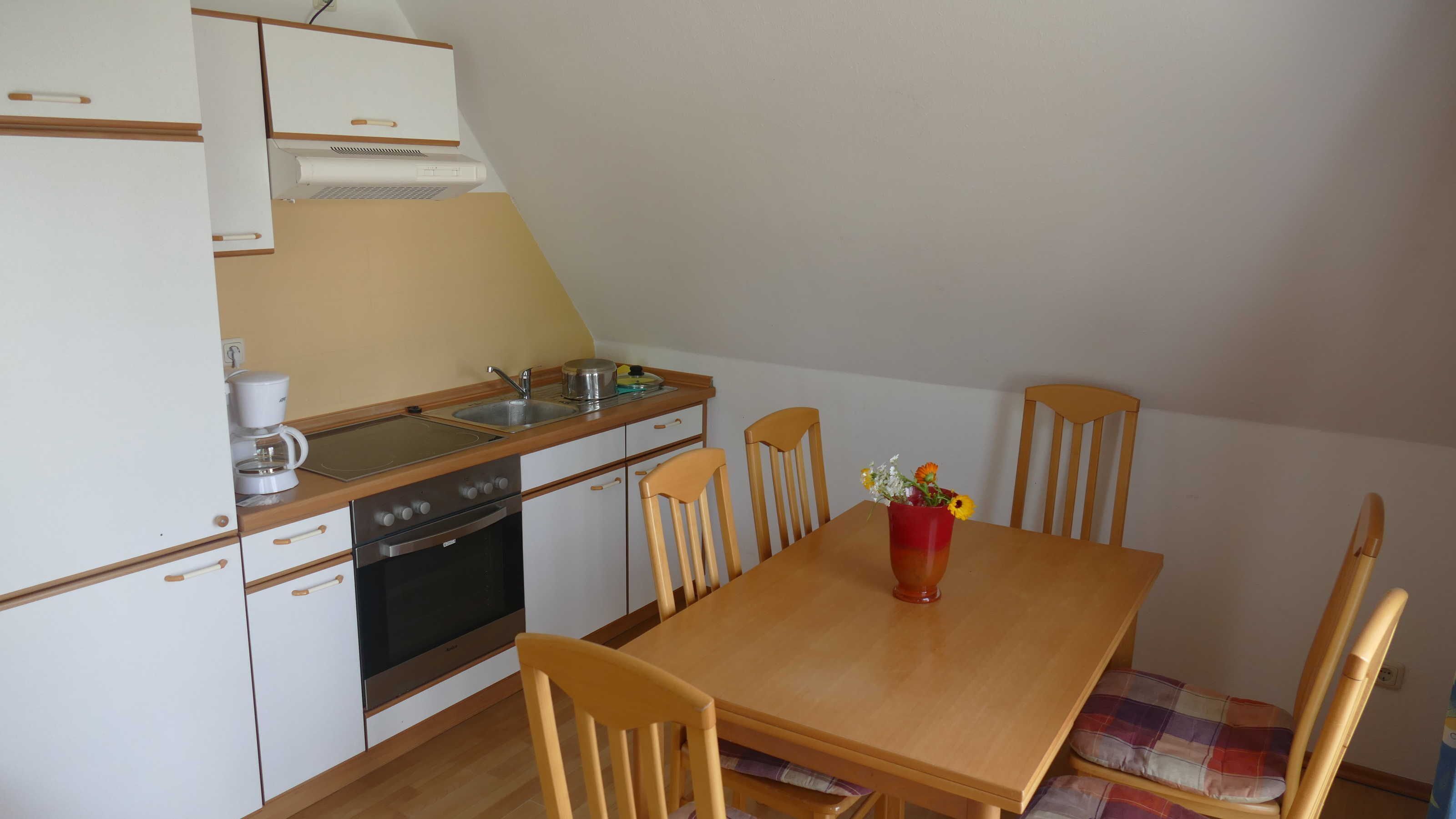 Una cocina compacta con estufa, horno, lavamanos y cafetera. Hay una mesa de comedor de madera con sillas y un florero con flores.