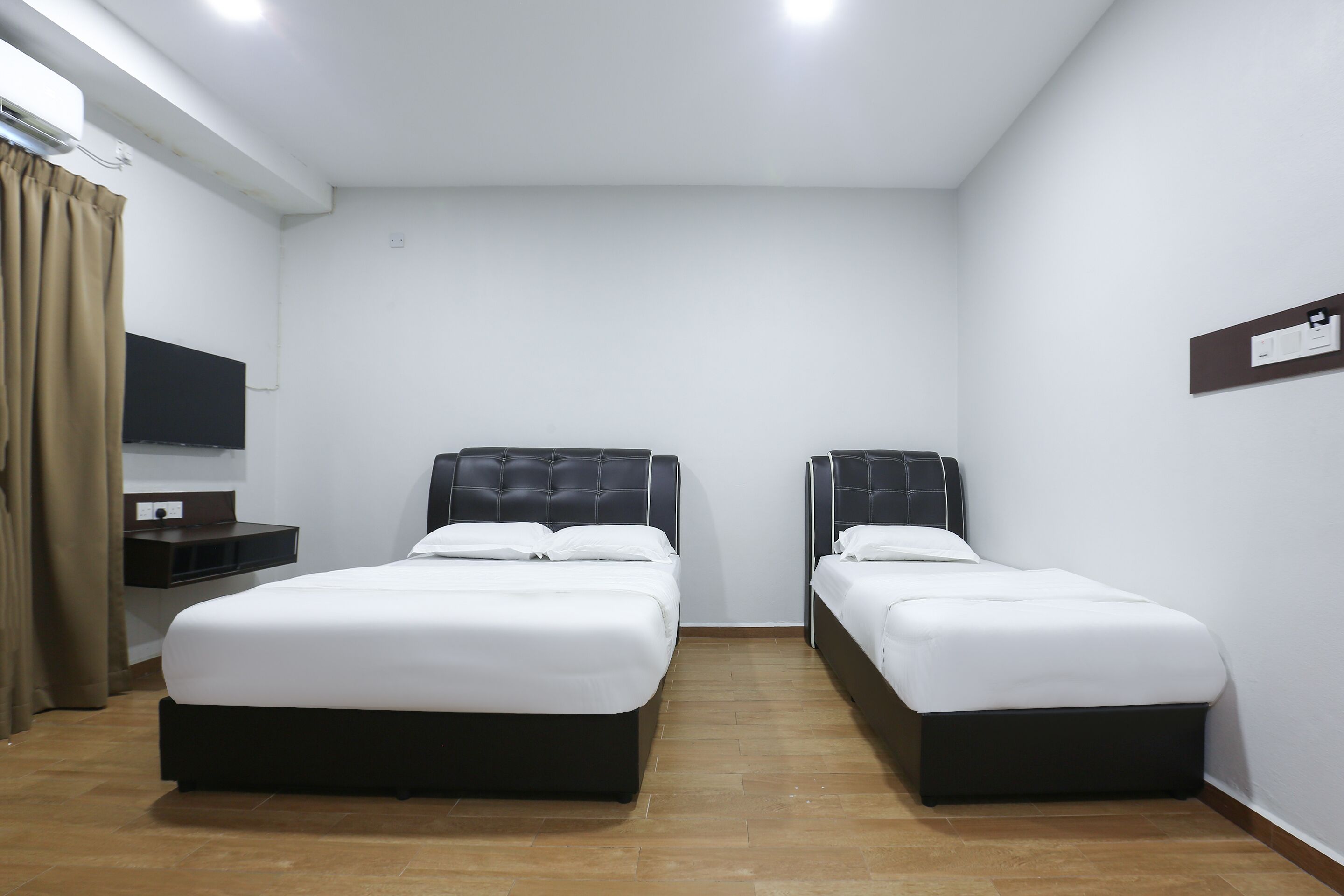 A hotel room with two single beds, a small bedside table, a flat-screen TV mounted on the wall, and a window with curtains.