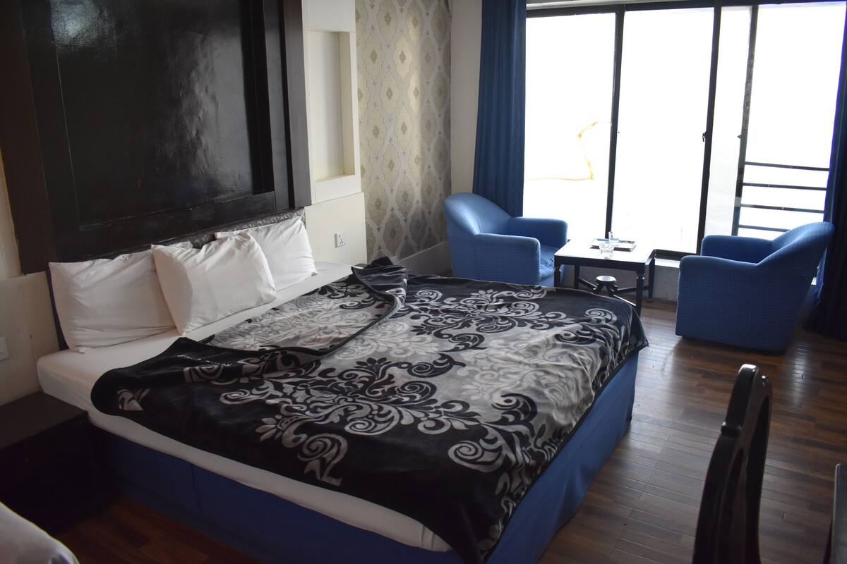 A hotel room with a bed, two blue armchairs, a small table, and a window with curtains.