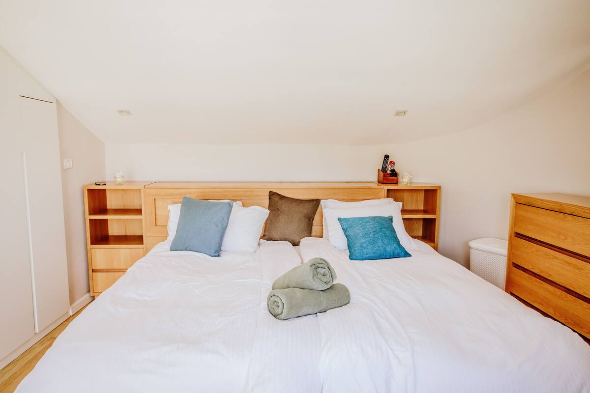 A neatly made bed with white linens and a pair of rolled towels. There are wooden headboards and a small shelf with a bottle and remote controls.