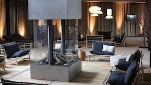 Et moderne loungeområde med en central pejs, behagelige siddepladser og et stort vindue med udsigt over naturen.