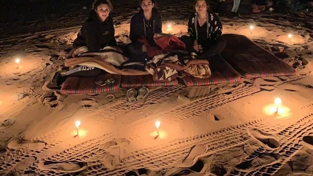 Three people sitting on a blanket with lit candles on a sandy beach at night. - Oryx camp (Wadi Rum)