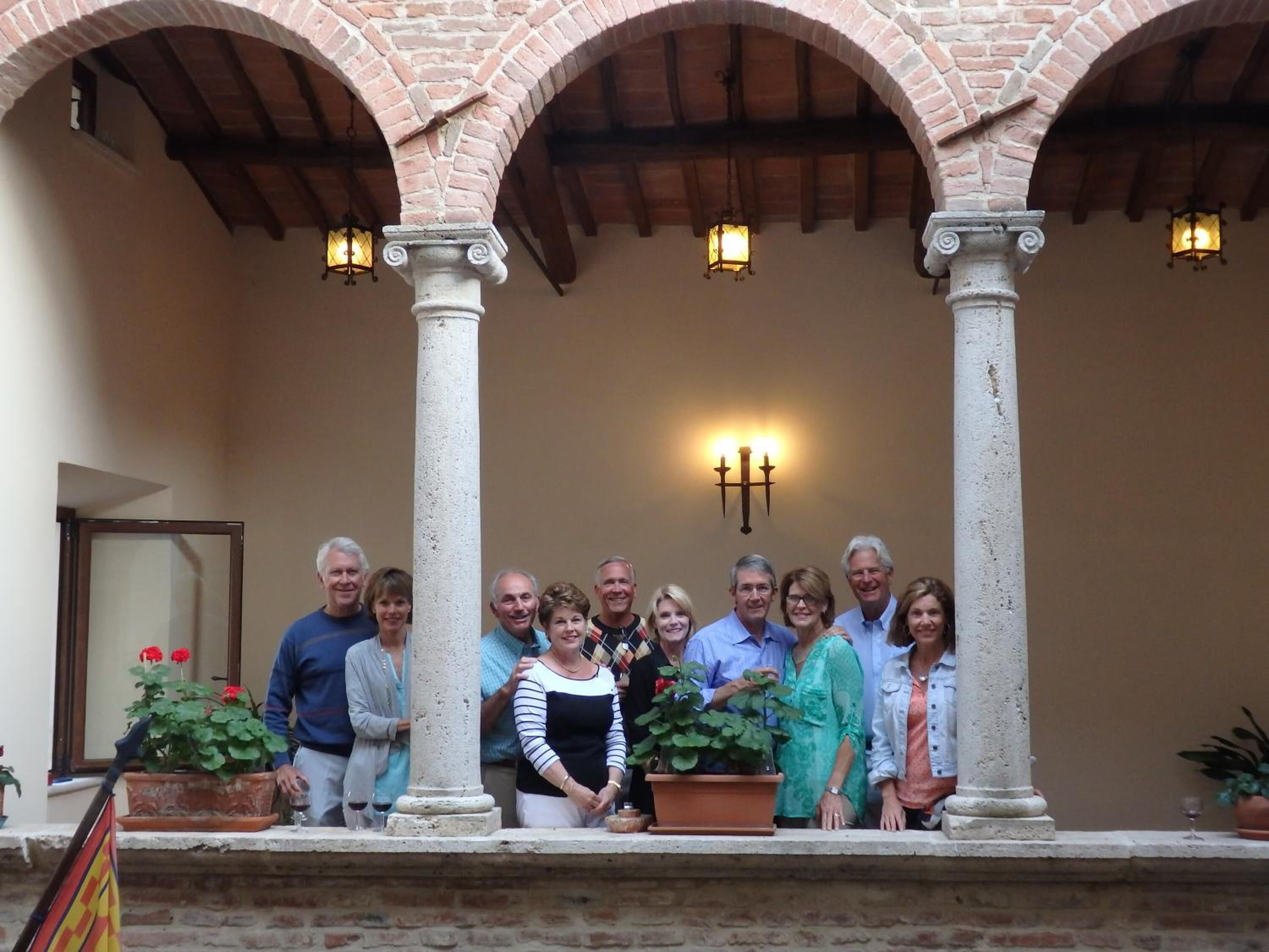 A group of people standing in a covered outdoor area with brick arches and columns, potted plants, and wall-mounted lighting.