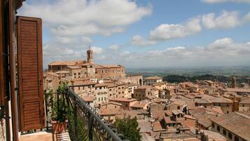 A view of a historic town with terracotta-roofed buildings, a prominent tower, and a balcony with a wooden shutter.