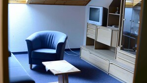 Une pièce avec un fauteuil bleu, un téléviseur posé sur un meuble et un buffet en bois doté de portes en verre.