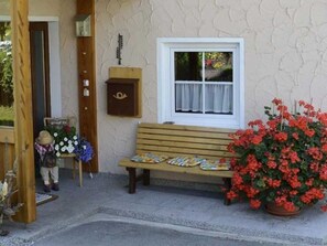 Eine Bank mit Kissen, eine Holzbank, ein Fenster mit Vorhängen, ein Blumentopf mit roten Blüten und eine Holztür.