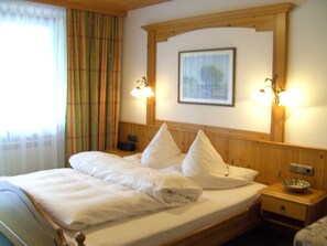 Ein Hotelzimmer mit einem hölzernen Kopfende, einem Bett mit weißen Leinen, zwei Nachttischlampen, einem gerahmten Bild an der Wand und einem Fenster mit Vorhängen.