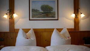 Une chambre d’hôtel avec une tête de lit en bois, deux lampes de chevet, un tableau représentant un paysage encadré et deux oreillers ornés d’une fleur rouge.