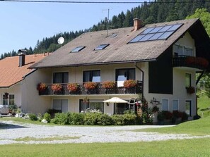 Ein zweistöckiges Gebäude mit geneigtem Dach, zahlreichen Fenstern und einem Balkon mit Pflanzgefäßen.