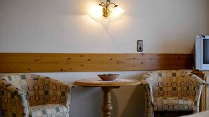 A room with two patterned chairs, a small round table with a bowl, a wall-mounted light fixture, and a television. - Double room 4 - Erd's Hoimatle (Jungholz)